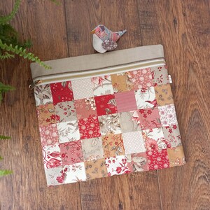 May include: A rectangular zipped pouch featuring a patchwork design of various floral and geometric fabric squares in shades of red, pink, beige, and white. The top section is a solid beige fabric with a metal zip, and a small bird ornament.