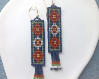 Op de afbeelding: Een paar kralen oorbellen met een rechthoekig ontwerp. De oorbellen hebben een marineblauwe achtergrond met een bloemenpatroon in oranje, wit en groen. Goudkleurige haken en kralenfranjes maken het ontwerp compleet.