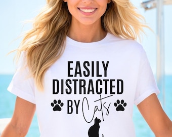 May include: White t-shirt with the text "EASILY DISTRACTED BY Cats" in black. The design includes paw prints and a black cat silhouette. The shirt is a casual style.