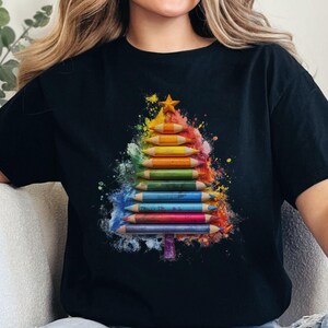 May include: Black t-shirt with a Christmas tree design made from coloured pencils. The tree is formed from a stack of pencils in various colours, topped with a gold star. The design has a watercolour effect.
