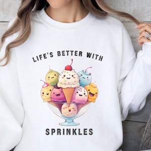 Op de afbeelding: Wit sweatshirt met een afbeelding van een kom ijs met sprinkels en de tekst "Life's Better With Sprinkles".