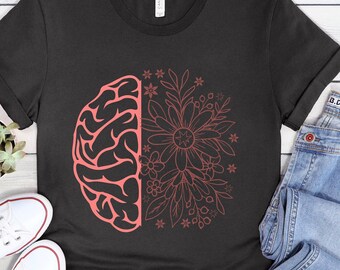May include: A black t-shirt with a pink outline of a brain and flowers on the front. The shirt is laid flat on a white wooden surface.