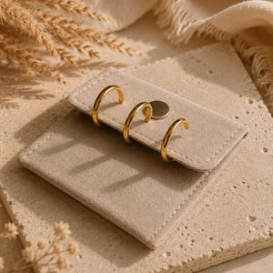 Könnte beinhalten: Ein quadratisches, beiges Schmuckdisplay aus samtartigem Material mit einem Klappverschluss, der durch einen runden Metalldruckknopf gesichert ist. Drei polierte goldfarbene Creolen sind am oberen Rand der Klappe in einer Reihe befestigt.