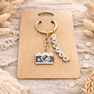 May include: A gold-toned metal keyring featuring a camera charm with white, black and gold enamel details. Attached to the split ring is a vertical strand of white cube beads with black lettering spelling out the name VERENA in capital letters.