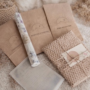 Könnte beinhalten: Eine Sammlung von Artikeln in Beigetönen, darunter Papierpakete mit Sonnen- und Bergillustrationen, eine floral gemusterte Kerze und ein verpacktes Paket, das mit Bindfaden verschnürt ist. Die Artikel sind auf einer weichen, strukturierten Oberfläche angeordnet.