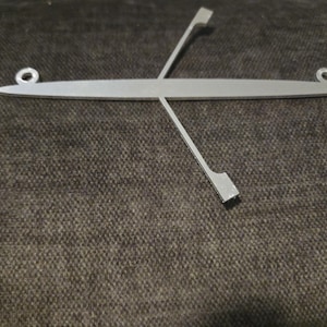 May include: A silver-colored, decorative metal rowboat with two oars. The rowboat has a long, slender body with a loop at each end for hanging. The oars are angled across the boat.