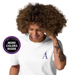 Camiseta con bordado delantero de letra A lila en forma de casa y chimenea. Camiseta algodón orgánico, unisex. Disponible en blanco y negro.