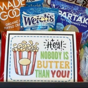May include: A gift basket filled with various snacks, including bags of puffed snacks, fruit snacks, and cookies. A sign with a popcorn graphic reads "Hey! Nobody is Butter Than You!" in colorful text.