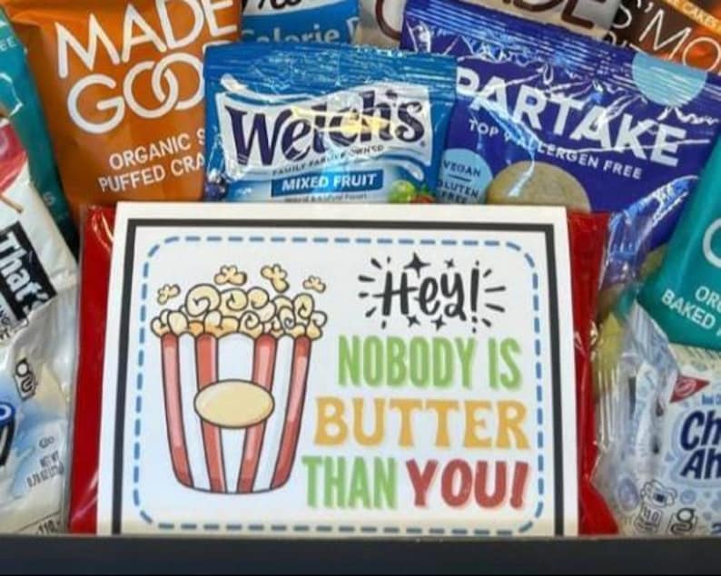 May include: A gift basket filled with various snacks, including bags of puffed snacks, fruit snacks, and cookies. A sign with a popcorn graphic reads "Hey! Nobody is Butter Than You!" in colorful text.