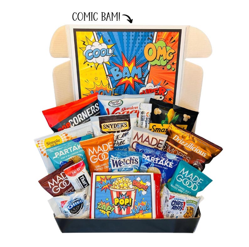 May include: A gift basket filled with various snacks, including chips, cookies, popcorn, and candy. The box features a comic book theme with bold colors and text like "COOL," "BAM," and "POP!" The snacks are arranged in a black tray.