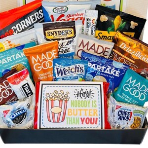 May include: A gift basket filled with various snack-sized food items, including popcorn, chips, cookies, and fruit snacks. The packaging features bright colors and brand names. A sign in the center reads "Hey! Nobody is Butter Than You!"