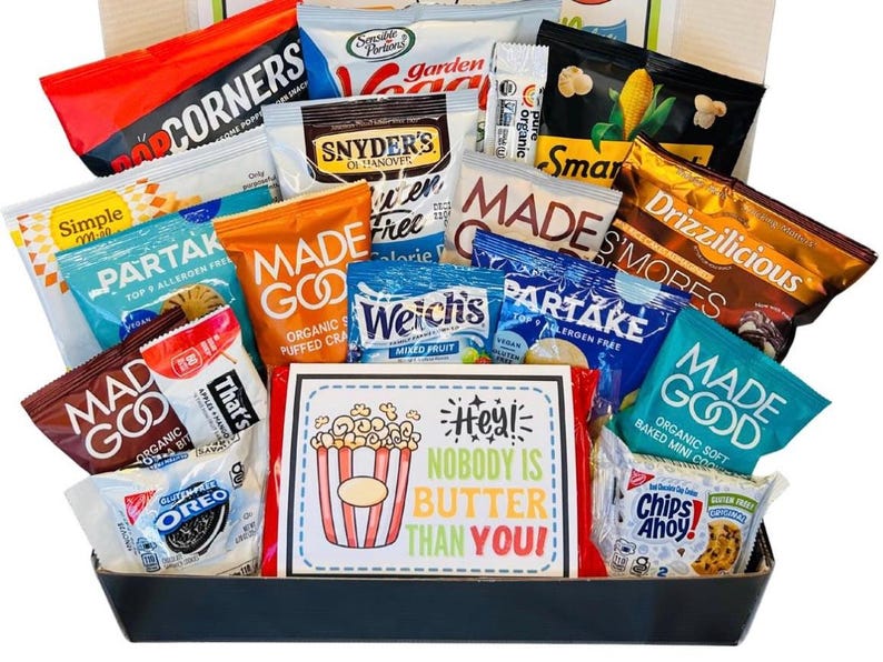 May include: A gift basket filled with various snack-sized food items, including popcorn, chips, cookies, and fruit snacks. The packaging features bright colors and brand names. A sign in the center reads "Hey! Nobody is Butter Than You!"