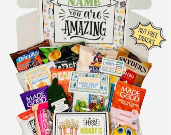 May include: A white gift box filled with assorted snacks, including Drizzilicious, Snyder's pretzels, and MadeGood cookie bites. It features cards with text like "YOU ARE AMAZING," "PLANT GOOD THOUGHTS," and "HEY! NOBODY IS BUTTER THAN YOU!" plus a small green crochet pickle.