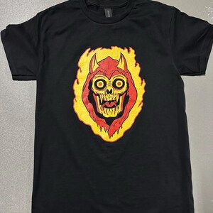 May include: Black t-shirt featuring a graphic of a red and yellow demon skull with horns and a fiery outline. The design has large yellow eyes and a menacing expression. The shirt is made of a soft material.