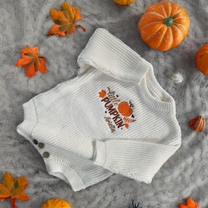 May include: Cream-colored baby sweater with the text "Little Pumpkin" and a pumpkin graphic. The sweater has three brown buttons and is surrounded by orange pumpkins and fall leaves on a gray textured surface.