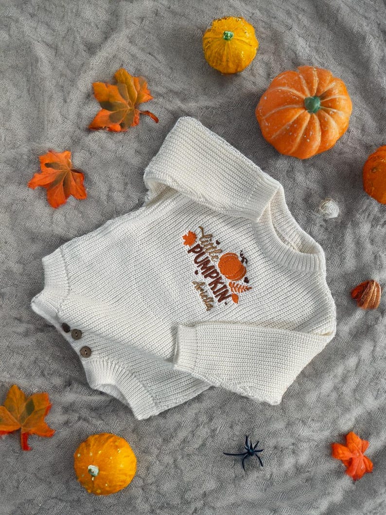 May include: Cream-colored baby sweater with the text "Little Pumpkin" and a pumpkin graphic. The sweater has three brown buttons and is surrounded by orange pumpkins and fall leaves on a gray textured surface.