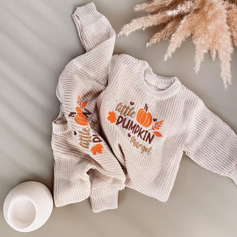 May include: A beige knit sweater with the words "little PUMPKIN margot" and a pumpkin graphic in orange and brown. The sweater has long sleeves and a ribbed hem and cuffs. The words "little" and a pumpkin graphic are also on the sleeve.
