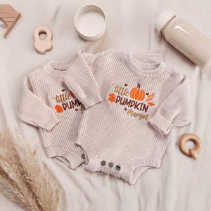May include: Two beige baby rompers with long sleeves and button closures. Each romper features the text "little PUMPKIN" with a pumpkin and leaf design. Additional wooden toys and a bottle are visible.