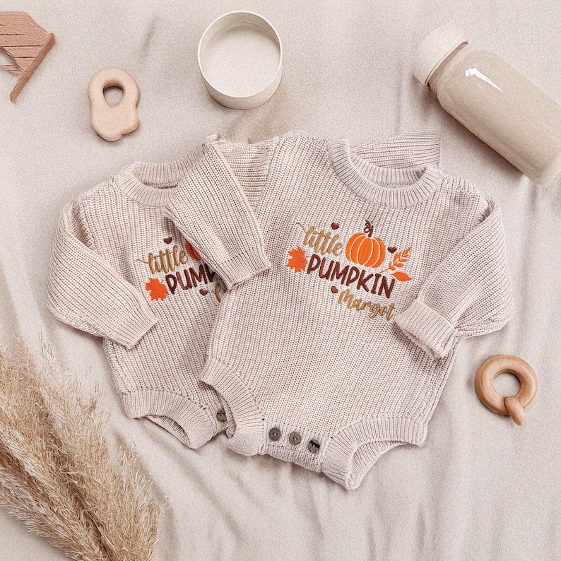 May include: Two beige baby rompers with long sleeves and button closures. Each romper features the text "little PUMPKIN" with a pumpkin and leaf design. Additional wooden toys and a bottle are visible.