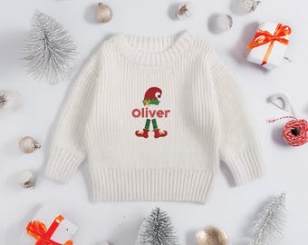 May include: A white knit sweater with the name "Oliver" and an elf graphic. The elf graphic includes a red hat, green collar, and green and red striped shoes. The sweater is surrounded by Christmas decorations.