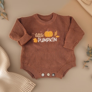 May include: A brown knit baby romper with long sleeves and three buttons at the bottom. The romper features the text "little PUMPKIN" with a pumpkin and leaf graphic in yellow and white.