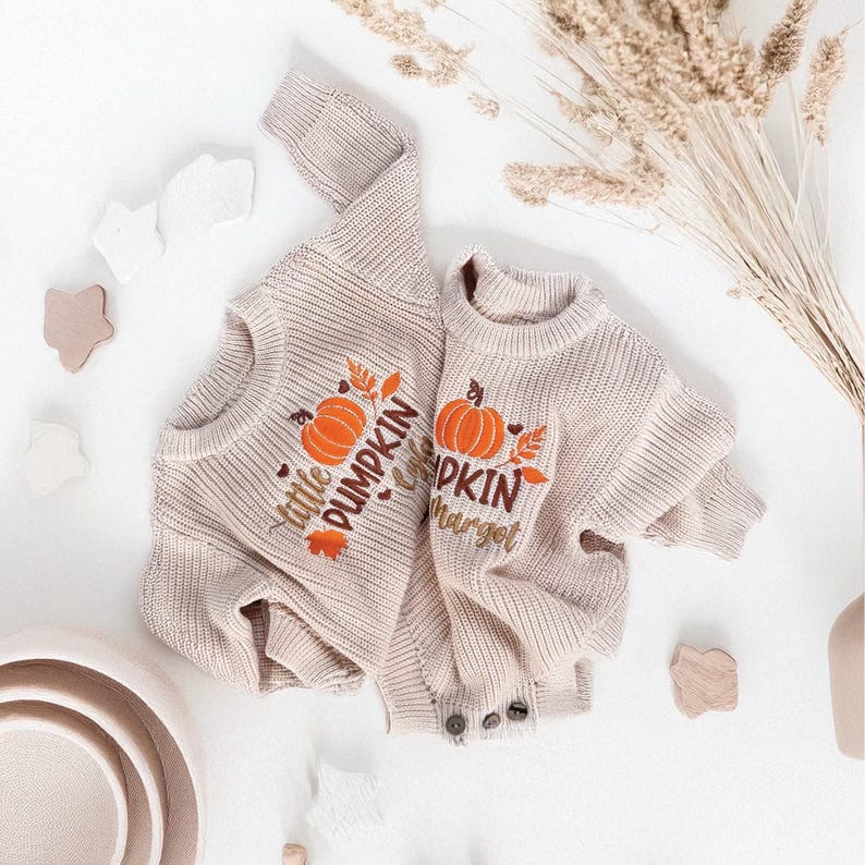 May include: Two beige knit sweaters, one adult and one child size, with orange pumpkin and leaf embroidery. The adult sweater says "Little Pumpkin" and the child's says "Pumpkin Hargel". The sweaters are on a white surface with decorative elements.