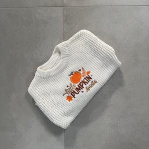 May include: White knit sweater with the embroidered text "Little Pumpkin Ariella" and a pumpkin graphic. The sweater is folded and lies on a gray tiled surface. The sweater is a fall-themed item.