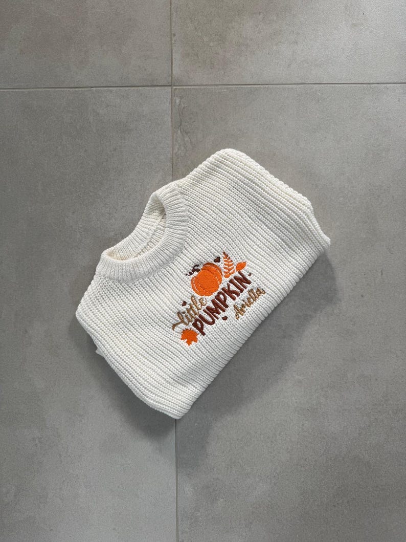May include: White knit sweater with the embroidered text "Little Pumpkin Ariella" and a pumpkin graphic. The sweater is folded and lies on a gray tiled surface. The sweater is a fall-themed item.