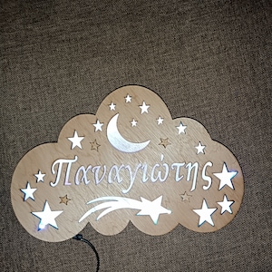 Peut inclure: Une veilleuse en bois en forme de nuage avec une lune croissante, des étoiles et le mot grec "Πανηγυριστής" (signifiant "célébrant"). La lumière émet une lueur blanche chaude.