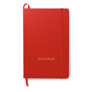 May include: A red notebook with a matching elastic band closure and a ribbon bookmark. The name "OLIVIA WILLIS" is printed in gold lettering on the front cover. The notebook is rectangular and has a textured surface.