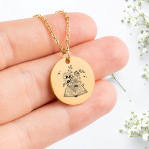 May include: Gold-colored necklace with a round pendant. The pendant features a black illustration of an open book with flowers and stars. The chain is delicate and the pendant is centered.