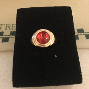 May include: A gold-toned ring with a large, round, red gemstone. The gemstone is set in a gold bezel with concentric circles. The ring is displayed on a black velvet surface, showcasing the jewelry's design and color.