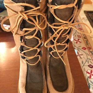 May include: A pair of brown suede boots with a lace-up design. The boots have a brown fur lining and a brown rubber sole.