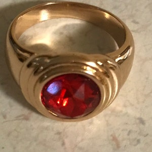 May include: A gold-colored ring with a large, oval-shaped, faceted red gemstone. The ring has a thick band and a raised setting for the gemstone. The gemstone reflects light, creating a sparkling effect.