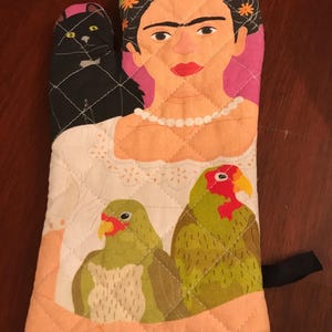 May include: A colorful oven mitt with a quilted design features a portrait of Frida Kahlo with a black cat, a pearl necklace, and two parrots.