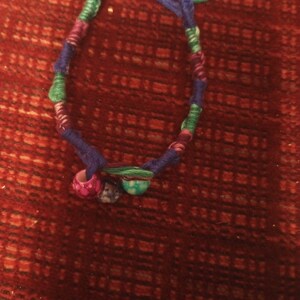 Puede incluir: Un brazalete de amistad multicolor con hilos azules, verdes, morados y blancos. El brazalete tiene dos pequeñas cuentas redondas con un patrón de mármol en tonos de rosa, azul y verde.