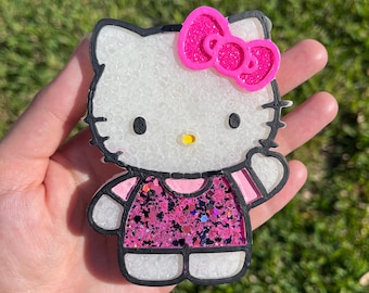 May include: A Hello Kitty-shaped decorative item with a white face, black outline, and a pink bow. The dress is filled with pink glitter. The item is held in a hand against a green background.