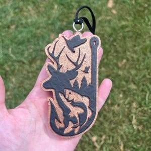 May include: A wooden ornament with a black silhouette of a deer, fish, and trees. The ornament is on a black cord and has a silver ring.