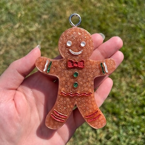 May include: A gingerbread man ornament with a silver loop for hanging. The ornament is brown with red, green, and white accents. It has a red bow tie, green buttons, and a smiling face. The ornament is held in a hand against a green background.