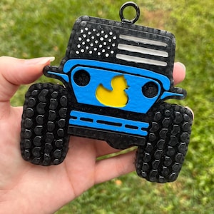 May include: A blue and black Jeep ornament with a yellow rubber ducky on the front. The Jeep has an American flag on the hood and black tires.