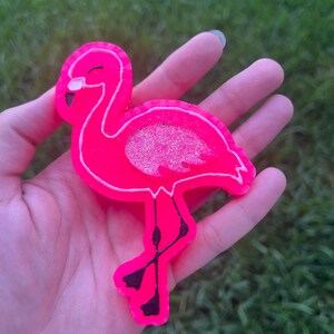 May include: A bright pink flamingo-shaped accessory with a white outline and glittery wing detail. The flamingo has black legs and a black beak. The accessory is held in a hand against a green background.