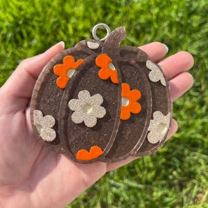 May include: A brown pumpkin-shaped keychain with orange and glittery white flower accents. The keychain has a silver loop for attachment. The design is set against a background of green grass.
