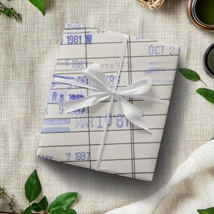 May include: A white gift wrapped in vintage style paper with a grid pattern and blue ink dates. The paper has dates from 1981 to 1987. The gift is tied with a white ribbon.