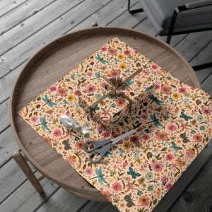 May include: A wrapped gift box and wrapping paper with a floral and insect pattern, including dragonflies and butterflies, are on a round wooden table. A pair of gray scissors and a silver ribbon are also on the table. The gift box is tied with a brown ribbon.