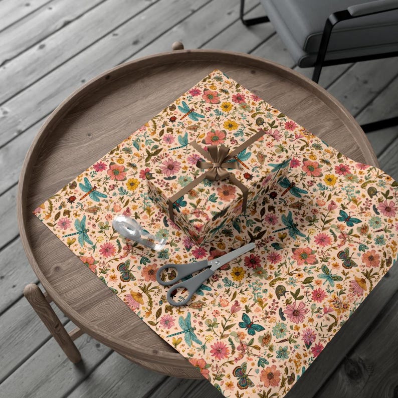 May include: A wrapped gift box and wrapping paper with a floral and insect pattern, including dragonflies and butterflies, are on a round wooden table. A pair of gray scissors and a silver ribbon are also on the table. The gift box is tied with a brown ribbon.