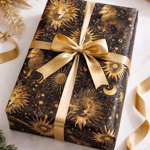 May include: A wrapped gift with black paper featuring gold lion illustrations, suns, moons, and stars. The gift is tied with a gold satin ribbon and bow. The overall theme is celestial and regal.