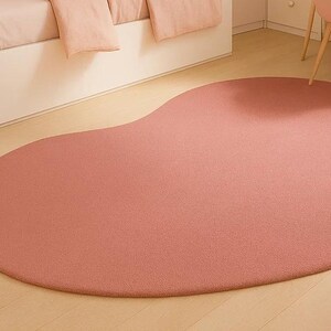 May include: A large, petal-shaped rug in a soft, muted rose color. The rug is made of a plush material and sits on a light wood floor. A stuffed elephant toy is visible in the corner, suggesting a children's room or nursery setting.