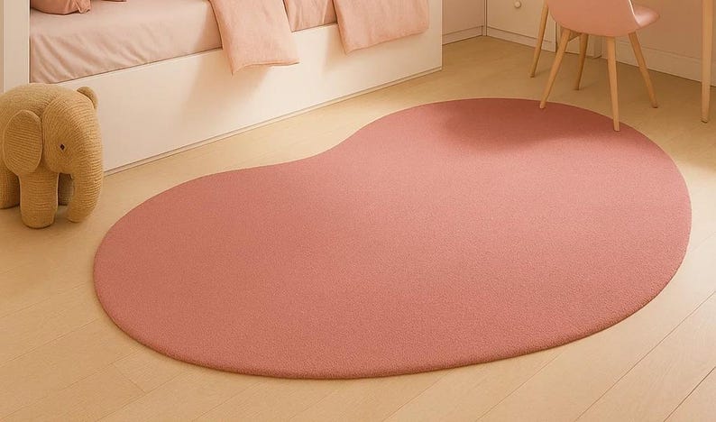 May include: A large, petal-shaped rug in a soft, muted rose color. The rug is made of a plush material and sits on a light wood floor. A stuffed elephant toy is visible in the corner, suggesting a children's room or nursery setting.