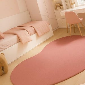 May include: A bedroom scene featuring a large, irregularly shaped, dusty rose rug. The room has a bed with matching bedding, a desk, and a chair. The walls and furniture are painted in soft, muted tones, creating a cohesive and calming aesthetic.