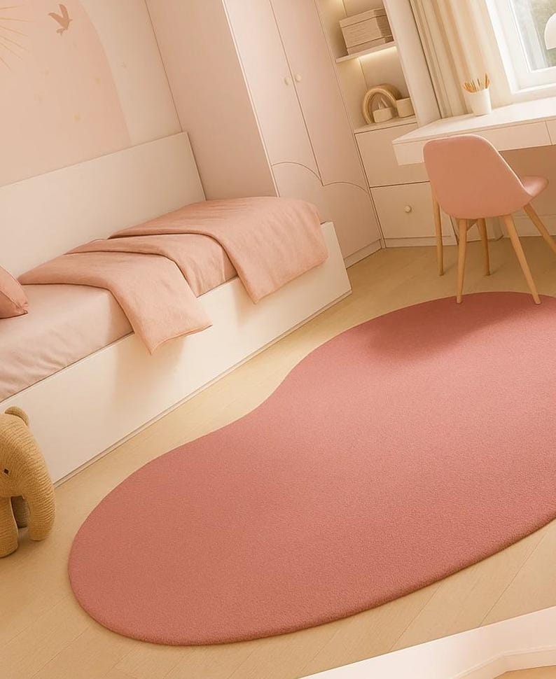 May include: A bedroom scene featuring a large, irregularly shaped, dusty rose rug. The room has a bed with matching bedding, a desk, and a chair. The walls and furniture are painted in soft, muted tones, creating a cohesive and calming aesthetic.